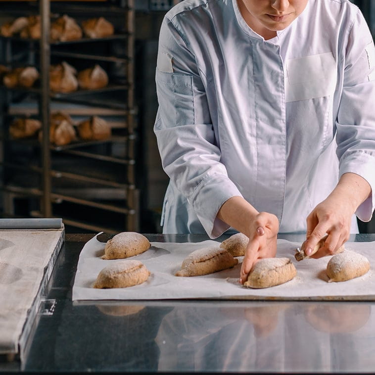 Bäckerin in weißer Bäckereibekleidung beim Einschneiden von Brötchenrohlingen auf Backblech in der Profi-Backstube